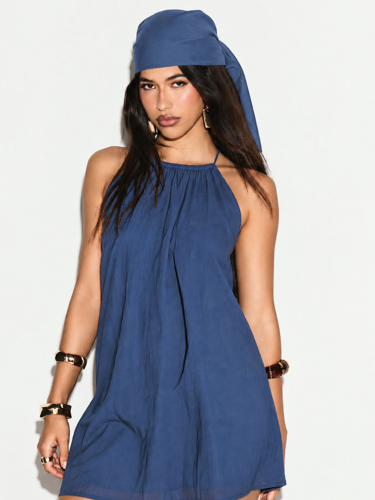 Front view of LILY blue halter neck mini dress with gathered neckline, worn with head scarf and gold bangles